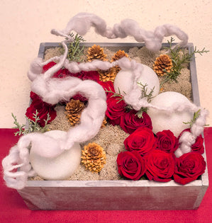 A small silver box brimming with sweetheart roses, spruce cones, reindeer moss, and juniper, accented with glittering white ornaments and silver yarn wrapped wire creating a snow flurry effect above the blooms.