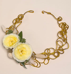 Gold aluminum wire that has been abstractly shaped to create the base of a necklace, adorned with white spray roses, fresh sage leaves, seeded eucalyptus, bunny tail grass, craspedia, and reindeer moss. 