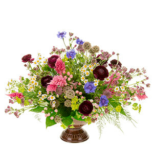 A rustic wildflower centerpiece in blue, dark burgundy, and pink colors mixes feverfew, scabiosa pods, bachelor buttons, bupleurum, plumosa, fatsia, Israeli ruscus, leather fern, and hypericum.