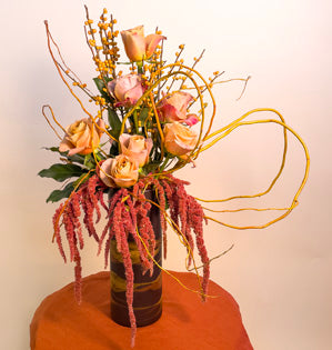A tall luxurious arrangement made with symbol roses, hanging amaranthus, curly willow, ilex berries, and fatsia leaves.