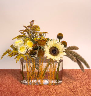 A horizontal textural arrangement created with sunflowers, solid aster, Princess Hoshi spray roses, banksia, cone flower, and millet, supported with an orange yarn armature.