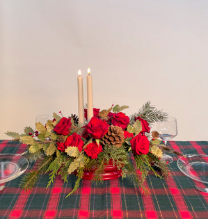 An oval crimson red vessel overflowing with a variety of evergreens, freedom roses, red carnations, and permanent botanical, topped off with two taper candles for a touch of romance.