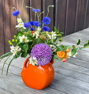 A vibrant Fiesta Ware pitcher made into a playful flower arrangement featuring spray roses, bachelor buttons, seasonal garden blooms.