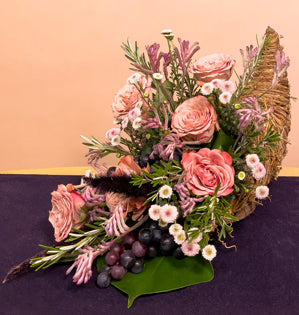 A bountiful cornucopia design full of aspidistra, monstera, rosemary, millet, kangaroo paw, Moccachino roses, spray chrysanthemums, and plastic grapes.