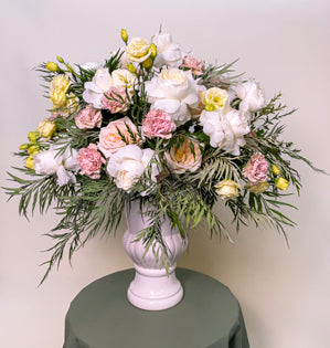 A large white urn overflowing with White Cloud and Princess Maya roses, antique carnations, yellow lisianthus, oregonia, and grevillea.