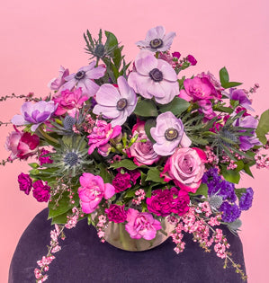 A bubble bowl vase full of Italian ruscus, salal, and fatsia leaves. With blooms of statice, leptospermum, eryngium, roses, mini carnations, freesia, and anemones all in shades of violet and fuchsia.