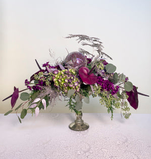 A contemporary horizontal design elevated by a mercury glass container, comprised of anthurium, permanent botanicals in a ruscus variety, hydrangea, both seeded and silver dollar eucalyptus, gilded plumosa, and stock, all in shades of violet.