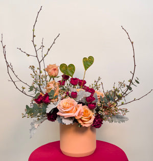A unique design featuring the Pantone color of the year - Peach Fuzz - paired with deep red hues made using dusty miller, eucalyptus, Carpe Diem roses, spray roses, carnations, wax flower and flowering quince, accented with galax leaf wired heart picks.