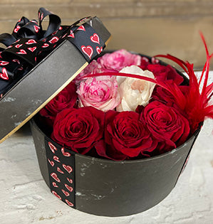 Red, pink, and cream colored roses rest in a black hat box decorated with pink heart-printed ribbon.