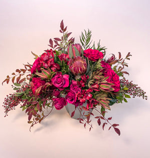 An exuberant hand-tied bouquet placed into a pale blue vase. Full of garden roses, spray roses leucadendron alstroemeria, protea, and pepperberry all in tints, tones, and shades of viva magenta.