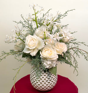 A lush white arrangement full winter blooms and foliage such as nerine lilies, O'Hara roses, hydrangea, and cypress.