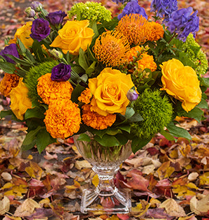 A vibrant round arrangement in a triadic color palette mixes Crème Brûlée roses, marigolds, lisianthus, green trick dianthus, miniature hydrangeas, asters, red huckleberry, and salal tips in a beautiful crystal vase.