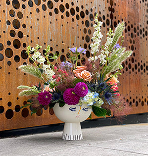 A unique ceramic vase with a face painted on is filled with summer blooms such as hydrangea, dahlia, roses, hypericum and mixed with smoke bush and a variety of grasses to add texture.