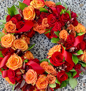 A beautiful sympathy flower wreath features High Magic orange roses and Freedom red roses mixed with leucadendron, myrtle, aspidistra leaves, and salal tips.