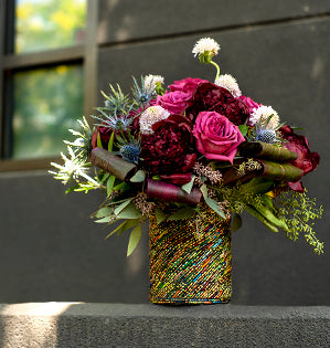 Peonies, roses, ti leaves, eryngium, scabiosa, Italian ruscus, Israeli ruscus, and seeded eucalyptus are combined in a glass vessel to create an eco-friendly bouquet of jewel-toned blooms.