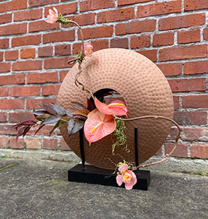 A bold modern round copper vessel features a pink anthurium nestled in the center, then adds twisted kiwi vine, miniature Cymbidium orchid blooms, leucothoe foliage, and a bit of preserved moss.