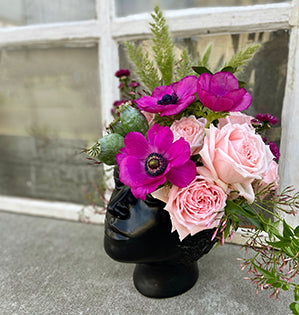 A unique head vase is filled with Princess Hitomi roses, Italian ruscus, anemone, poppy pods, bunny tail grass, mini aster, and jasmine.