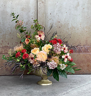 A bold bespoke design in a gold compote is filled with fatsia, huckleberry, Israeli ruscus, agonis, viburnum, roses, carnations, pin cushion protea, spray roses, blackberries, and butterfly ranunculus.