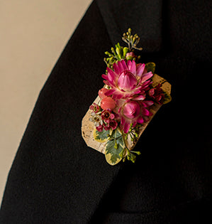 A small boutonniere uses pink strawflower, seeded eucalyptus, variegated pittosporum, wax flower, and hypericum berries, then adds a bark ribbon for a base.