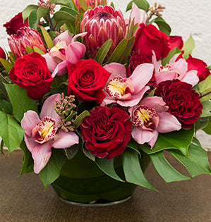 A unique and gorgeous Valentine’s Day floral design mixes Freedom roses, Hearts roses, cymbidium orchids, protea, aspidistra leaves, split-leaf philodendron, salal tips, and sumac.
