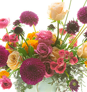 A vibrant and juicy-colored boho floral design mixes Italian ruscus, variegated pittosporum, acacia foliage, dahlias, marigolds, Blueberry roses, Shimmer roses, spray roses, ranunculus, and scabiosa in a pottery vessel.
