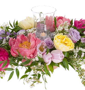 A soft and romantic wedding floral centerpiece mixes roses, peonies, lupine, tulips, spray roses, spirea, and Italian ruscus, then adds a glass hurricane with candle for an elegant effect.