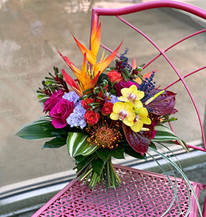 A vibrant hand-tied tropical bouquet is comprised of heliconia, anthurium, miniature Phalaenopsis orchids, pincushion protea, kangaroo paws, lavender, and roses.