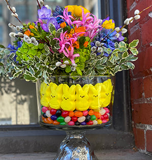 This vibrant, beautiful Easter floral design consists of a hand-tied bouquet that mixes ranunculus, nerines, freesia, hyacinth, hydrangea, dubium, and pussy willow set into a compote bowl lined with bright colored Peeps candy and jelly beans.