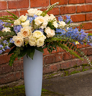 A tall vase arrangement features garden roses, blooming branches, delphiniums, and eucalyptus for a high-impact floral design.