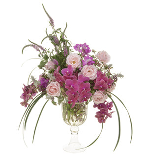 A tall floral centerpiece has elegant draping phalaenopsis orchids, soft pink garden roses, delicate mint and oregano, yarrow, hot pink freesia, towering veronica, lamb's ear, and lily grass in an elevated glass vase. 