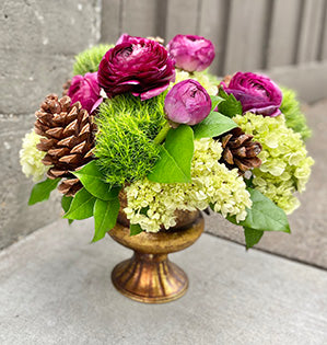 A compote arrangement in green, yellow and purple colors is filled with hydrangeas, dianthus, ranunculus, and salal tips.