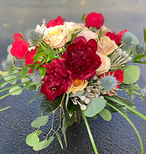 A wedding bouquet mixes several kinds of garden roses in red and blush colors with peonies, eryngium, berzillia, silver dollar eucalyptus, fatsia leaves, lily grass, and Italian ruscus.