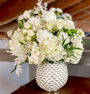 A floral arrangement in white mixes hydrangeas, mini carnations, monte casino, and spray roses with Italian ruscus, salal, and aspidistra.