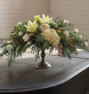 Floral arrangement on a pedestal with greenery and white flowers on a dark surface.