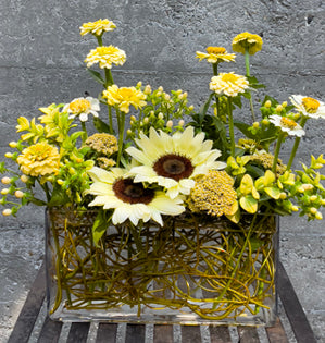 Decorative arrangement of sunflowers and other flowers in a woven basket on a gray surface.