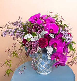 A lovely garden style design in a blue vase made with, lilacs, phalaenopsis orchids, carnations, dusty miller, and jasmine vine.