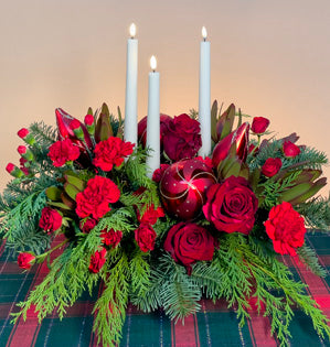 Decorative floral arrangement with red flowers, greenery, and white candles on a plaid tablecloth.
