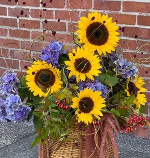 Bouquet of sunflowers and purple flowers in a woven basket against a brick wall.