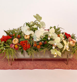 A long rectangular floral arrangement in a light-colored planter with gold-tone legs, featuring white orchids, red and orange flowers, and green foliage, set against a plain light background.