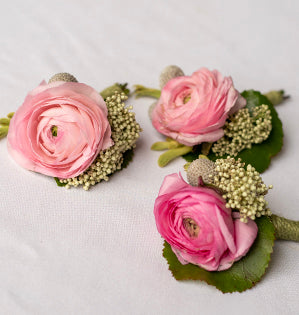 Three pink ranunculus wrist flowers with green leaves and small white filler blooms, arranged on a white fabric background.