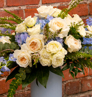 Bouquet of white and light blue flowers in a vase against a brick wall.